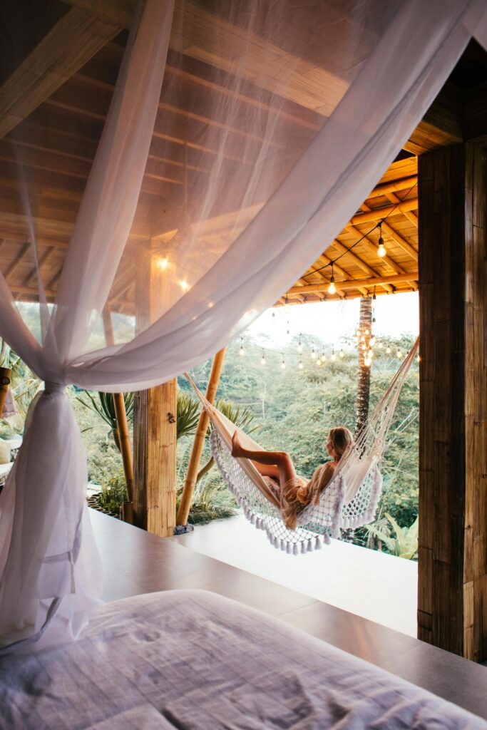 Side view of unrecognizable barefoot female traveler lying in hammock on bamboo terrace against bed under canopy